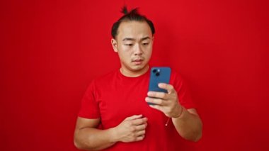 Üzgün genç Çinli bir adam, kırmızı arka planda izole edilmiş, akıllı telefondan mesaj atıyor, bir sorun için endişeleniyor.