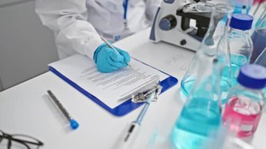 Çekici, sarışın, genç bir bilim adamının ilginç portresi, tıbbi araştırmalara dalmış, laboratuarda mikroskop altında incelerken tutkuyla not alıyor.