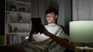 Genç İspanyol ergen rahat bir şekilde yatağında otururken gülümsüyor. Sıcak yatak odasında, dokunmatik bir ped kullanarak günün tadını çıkarıyor.