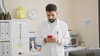 Sakallı İspanyol adam, laboratuvarda beyaz laboratuvar önlüklü akıllı telefon kullanırken gülümsüyor..