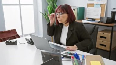Olgun bir kadın, etrafı teknoloji ve ofis malzemeleriyle çevrili iyi aydınlatılmış ofisinde özenle çalışır..