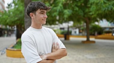 Neşeli genç İspanyol genç, kendine güvenle ışıldıyor, neşeli bir kolla neşe ifade ediyor yemyeşil bir şehir parkının kalbinde çapraz bir jest yapıyor.