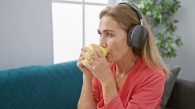 Orta yaşlı bir kadın oturma odasında kahve içerken kulaklıkla müzik dinliyor.