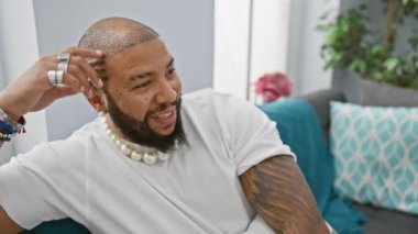 Güler yüzlü, sakallı, dövmeli, inci takan, evin içinde gülen siyah bir adam.