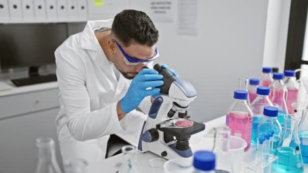 カラフルな試験管とフラスコに囲まれた科学研究室の顕微鏡を使用した実験室のコートの男 — ストック動画