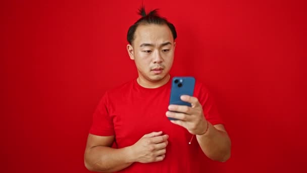 Расстроенный Молодой Китаец Изолированный Красном Фоне Пишет Смс Смартфоне Беспокоится — стоковое видео