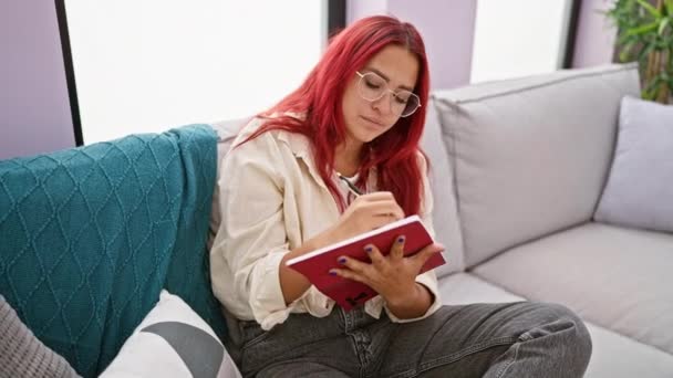 严肃的红头发女人 舒服地坐在沙发上 全神贯注地在一本书里写着便条 陷入沉思 在家里房间里悠闲的氛围中 — 图库视频影像