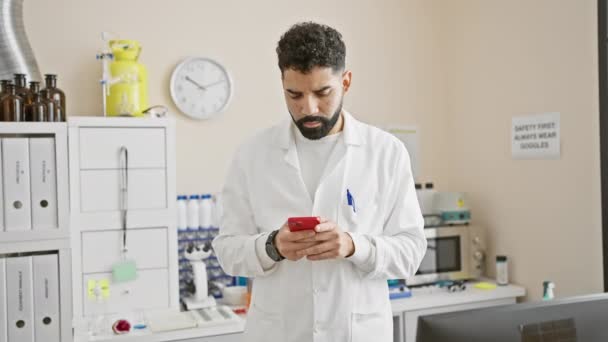 ヒスパニックのヒスパニック男性は 医学研究室で屋内で白い実験室のコートでスマートフォンを使用しながら微笑みます — ストック動画