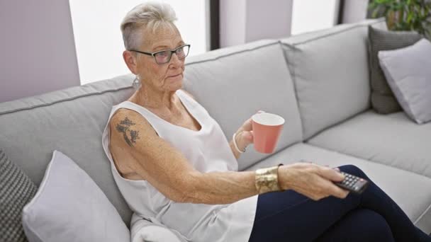シニアの灰色の髪の女性は 暖かいコーヒーのカップとソファーに座って テレビで彼女のお気に入りの映画を見てリラックス — ストック動画