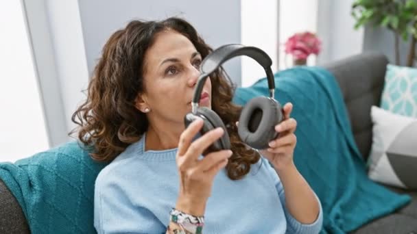 カーリーヘアのある中年の女性は 屋内ソファでリラックスしてヘッドフォンで音楽を楽しんでいます — ストック動画