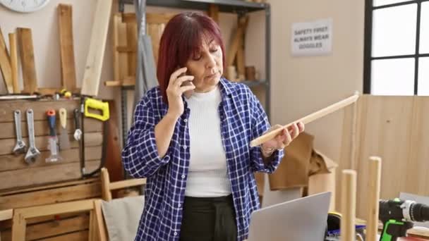 成熟した女性が見えるツールとラップトップを備えた大工のワークショップで電話で話しながら木片を検査します — ストック動画