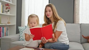 Ciddi beyaz anne ve küçük kızı evde oyuncaklarla çevrili bir kanepede oturup kitap okumanın keyfini çıkarıyorlar.