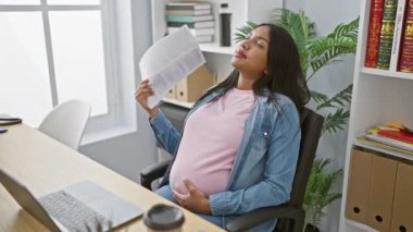Genç hamile iş kadını, sıcak ofisten gelen rahatsızlıkla boğuşuyor, belgeyi el hayranı olarak kullanırken iş talebine dalıyor..