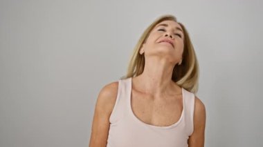 Olgun sarışın bir kadın, beyaz bir duvara karşı güven içinde poz verir. Öylesine bir ortamda zarafet ve güzellik yayar..