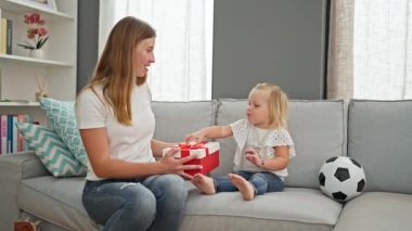 Beyaz bir anne ve kızın evde birlikte bir doğum günü hediyesi açması, sıcak gülümsemeleri odayı aydınlatıyor.