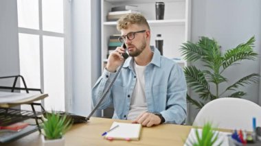 Mavi gözlü ve sakallı genç bir beyaz adam modern ofis ortamında telefonla konuşuyor..