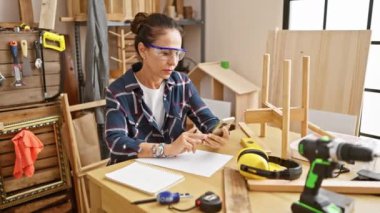 Orta yaşlı kıvırcık saçlı İspanyol bir kadın marangozluk atölyesinde aletler ve ahşaplarla dolu akıllı telefonuna odaklanmış..