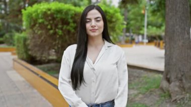 Kendine güvenen İspanyol bir kadın, sakin bir şehir parkında kollarını kavuşturup gülümsüyor.,