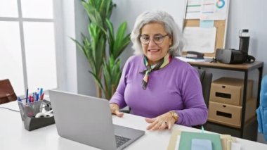 Orta yaşlı bir kadın modern ofis ortamında video aracılığıyla iletişim kuruyor, profesyonellik ve nişan gösteriyor.