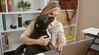 Genç sarışın bir kadın modern ofis ortamında siyah labrador köpeğini kucaklıyor, sıradan bir çalışma ortamını yansıtıyor..