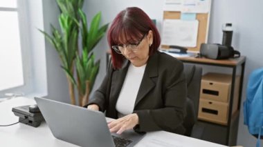 Orta yaşlı İspanyol kadın dizüstü bilgisayarla modern ofis ortamında çalışıyor, profesyonellik ve konsantrasyon yayıyor..
