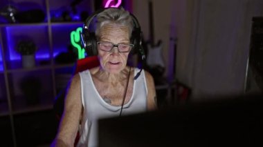 Kendine güvenen, gri saçlı, son sınıf öğrencisi, gece oyun odasında fütüristik video oyunu oynarken gülümseyen ve modern teknolojiyle eğlenen bir kadın.