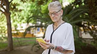 Havalı, gri saçlı son sınıf öğrencisi kadın ciddi şekilde internetten mesajlaşmaya odaklanmış. Güneşli park yeşilliğinin ortasında duruyor. Teknoloji meraklısı olgun bir yaşam tarzı sergiliyor..