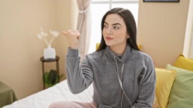 Neşeli, genç, İspanyol bir kadın pijama giymiş, güzel bir şekilde yatak odasında oturuyor, başparmağını kaldırmış ve başka tarafa bakıyor, ve tüm bunlar olurken güzel, parlak bir gülümsemesi var.