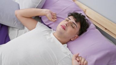Genç bir adam, huzurlu bir yatak odasında mor yatak örtüsüyle çevrili huzurlu bir ifadeyle yatakta dinleniyor..