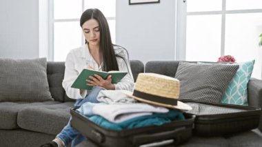 Genç bir kadın oturma odasında bir kanepede oturuyor, yanında bir bavul ve şapkayla bir deftere yazıyor..