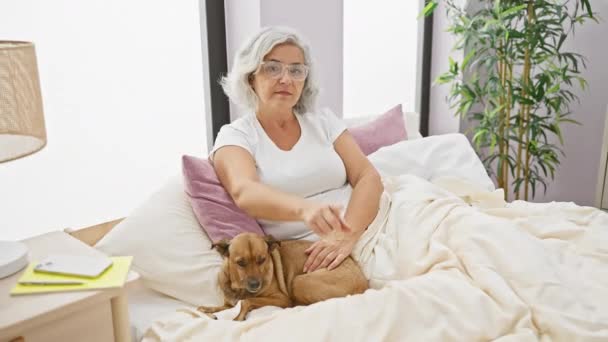自信的 灰白头发的中年妇女 穿着睡衣 从床上指着你 狗肩并肩地看着你 严肃地看着你的脸 卧室里 — 图库视频影像
