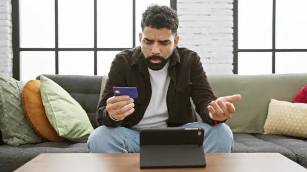 头戴胡子的困惑男人坐在沙发上 凝视着客厅里的平板电脑 手里拿着信用卡 — 图库视频影像