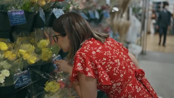 メガネ付きの豪華なヒスパニック女性は 自信を持って立っている間に喜びを放射し 晴れた都市のショッピングセンターでカラフルな花を嗅ぎます — ストック動画