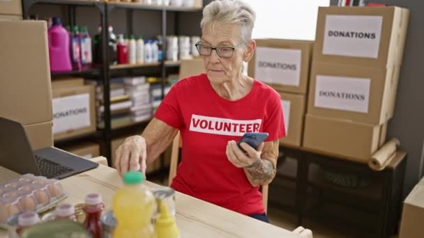 在慈善中心的利他主义活动中 一位高个子 头发灰白 身穿眼镜的女志愿者巧妙地使用她的智能手机检查装满捐赠产品的盒子 — 图库视频影像