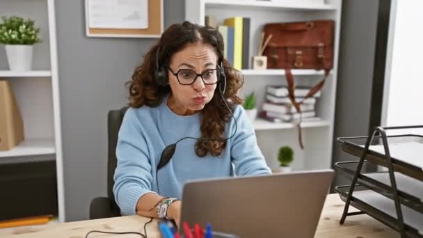 一位头发卷曲 头戴耳机的中年惊慌失措的女人心满意足地在办公室的办公桌前工作 — 图库视频影像