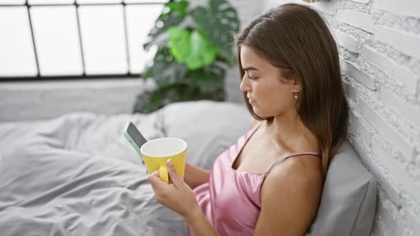 迷人的年轻惊慌失措的女人穿着睡衣坐在舒适的床上 静静地喝着早茶 同时在舒适的卧室里用智能手机发短信 — 图库视频影像