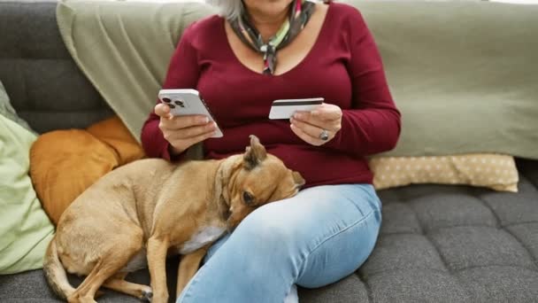 オンライン決済のためにスマートフォンとクレジットカードを使用して成熟した女性 屋内ソファの彼女の横に彼女の犬と — ストック動画