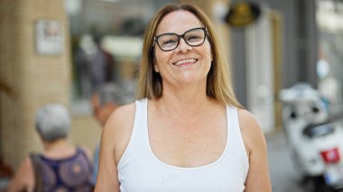 Orta yaşlı İspanyol kadın gözlüklü, sokakta gülümsüyor.