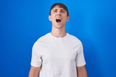Kafkasyalı sarışın adam mavi arka planda kızgın ve kızgın bağırıyor sinirli ve öfkeli, öfkeyle bağırıyor. öfke ve agresif konsept. 
