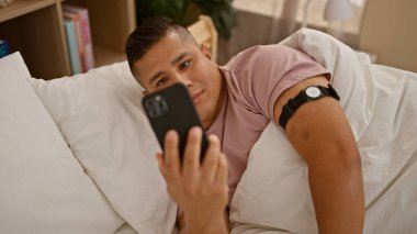 Young latin man using smartphone lying on bed at bedroom