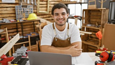 İyi donanımlı bir marangozhanede gülümseyen dizüstü bilgisayarlı sakallı bir adam..