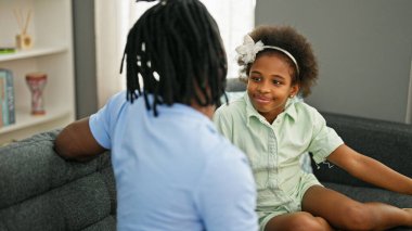 Afro-Amerikan baba ve kızı gülümsüyor. Kanepede oturuyorlar ve evde konuşuyorlar.