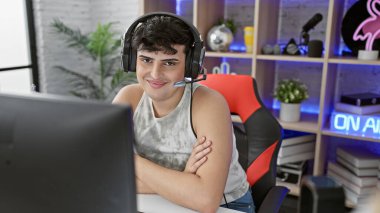 Modern oyun odasında kulaklığı olan genç adam masaya yaslanmış gülümsüyor.,