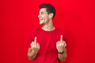 Young hispanic man standing over red background showing middle finger doing fuck you bad expression, provocation and rude attitude. screaming excited 