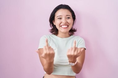Hispanic young woman standing over pink background showing middle finger doing fuck you bad expression, provocation and rude attitude. screaming excited 