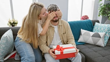Bir kadın, hoş bir oturma odasında hediyeyle bir erkeği şaşırtır. Ev ortamında aşkı ve birlikteliği sembolize eder..
