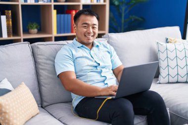 Dizüstü bilgisayar ve kulaklık kullanan genç Çinli adam evdeki kanepede oturuyor.