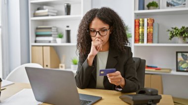 Genç ve güzel İspanyol iş kadını. Ofiste dizüstü bilgisayar ve kredi kartı kullanıyor.