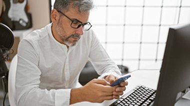 Radyo stüdyosunun kalbinde, gri saçlı, genç İspanyol adam yoğun bir şekilde canlı yayında haberleri sunuyor, akıllı telefondan hayati bilgileri mesaj atarken ciddi bir yüz ifadesine sahip.