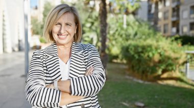 Şehir parkında kollarını kavuşturmuş kendinden emin, orta yaşlı sarışın bir kadın..